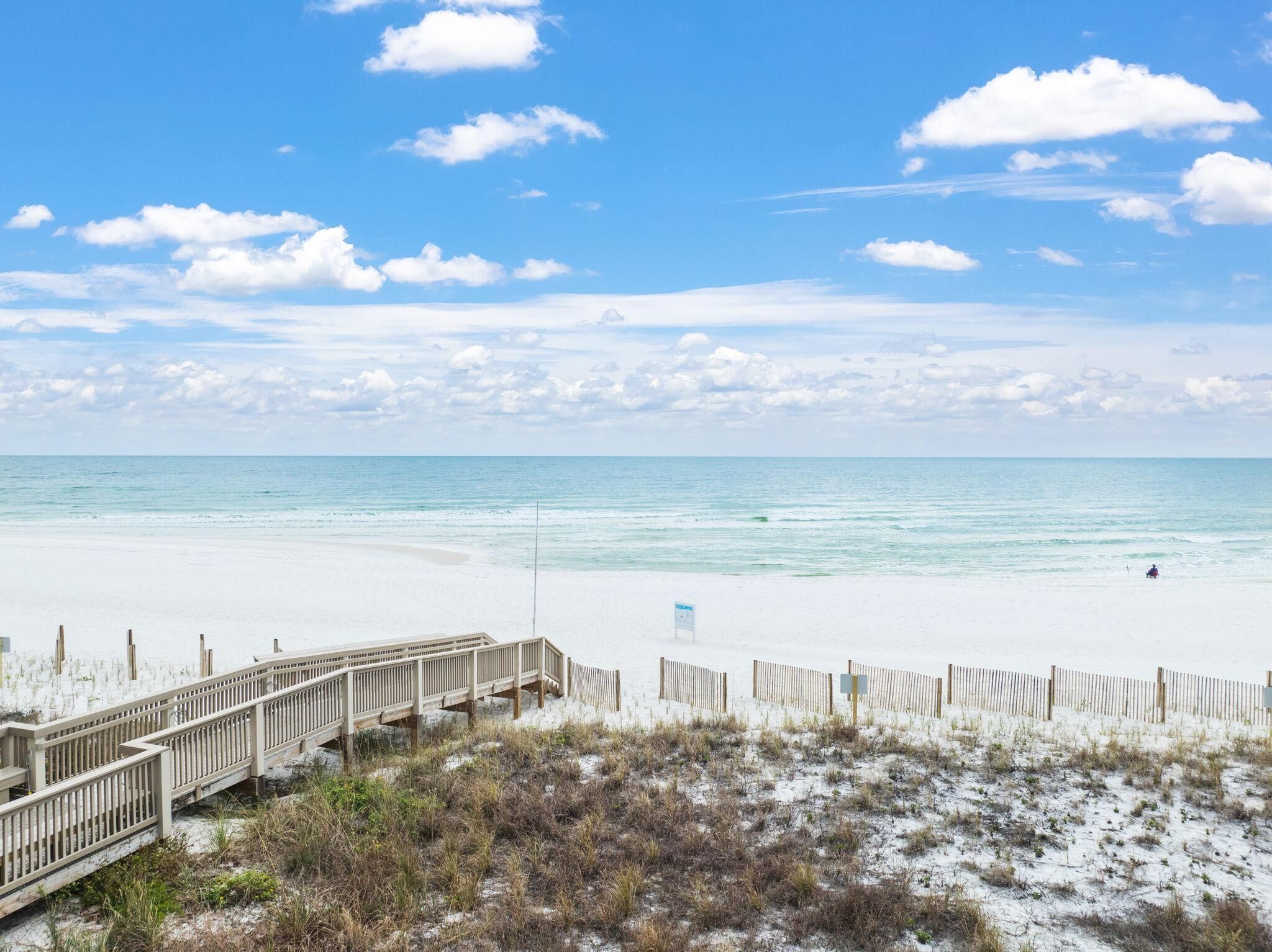 A beach with a wooden boardwalk leading to the shore, sandy dunes, and a clear blue sky.