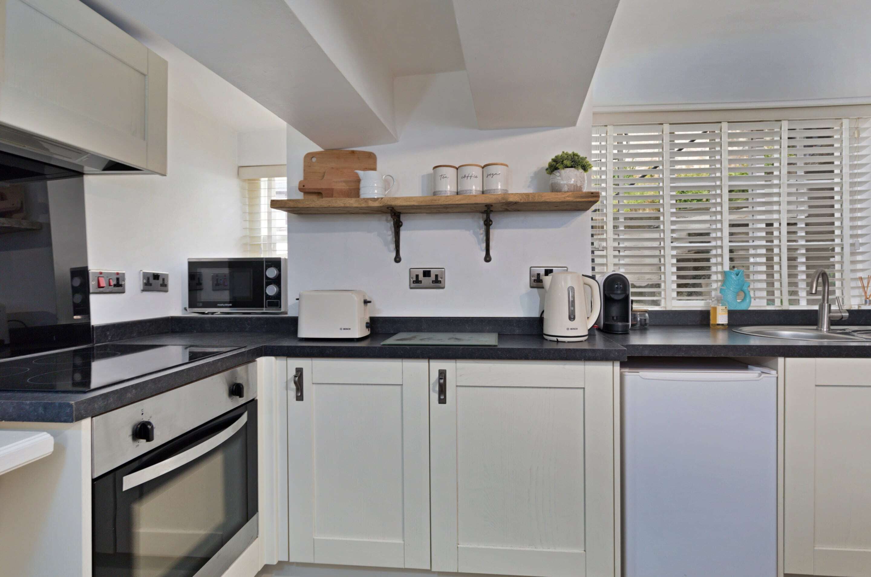 A kitchen with white cabinets, a black countertop, a microwave, a toaster, a coffee maker, and a window with blinds.