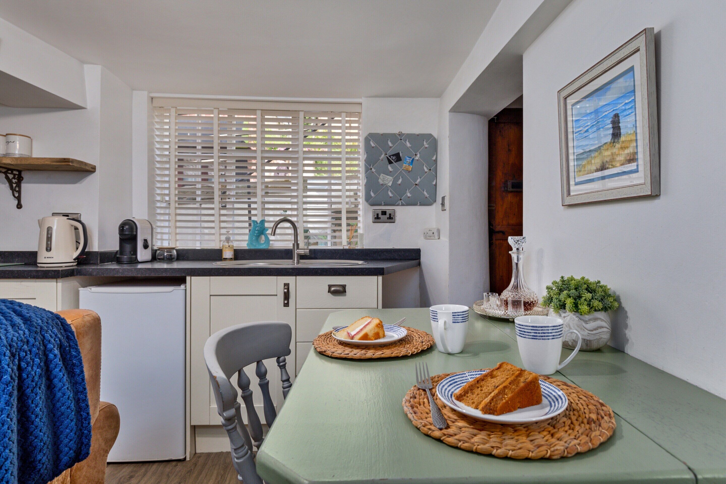 A compact kitchen with a dining area, a small refrigerator, a coffee maker, and a window with blinds.
