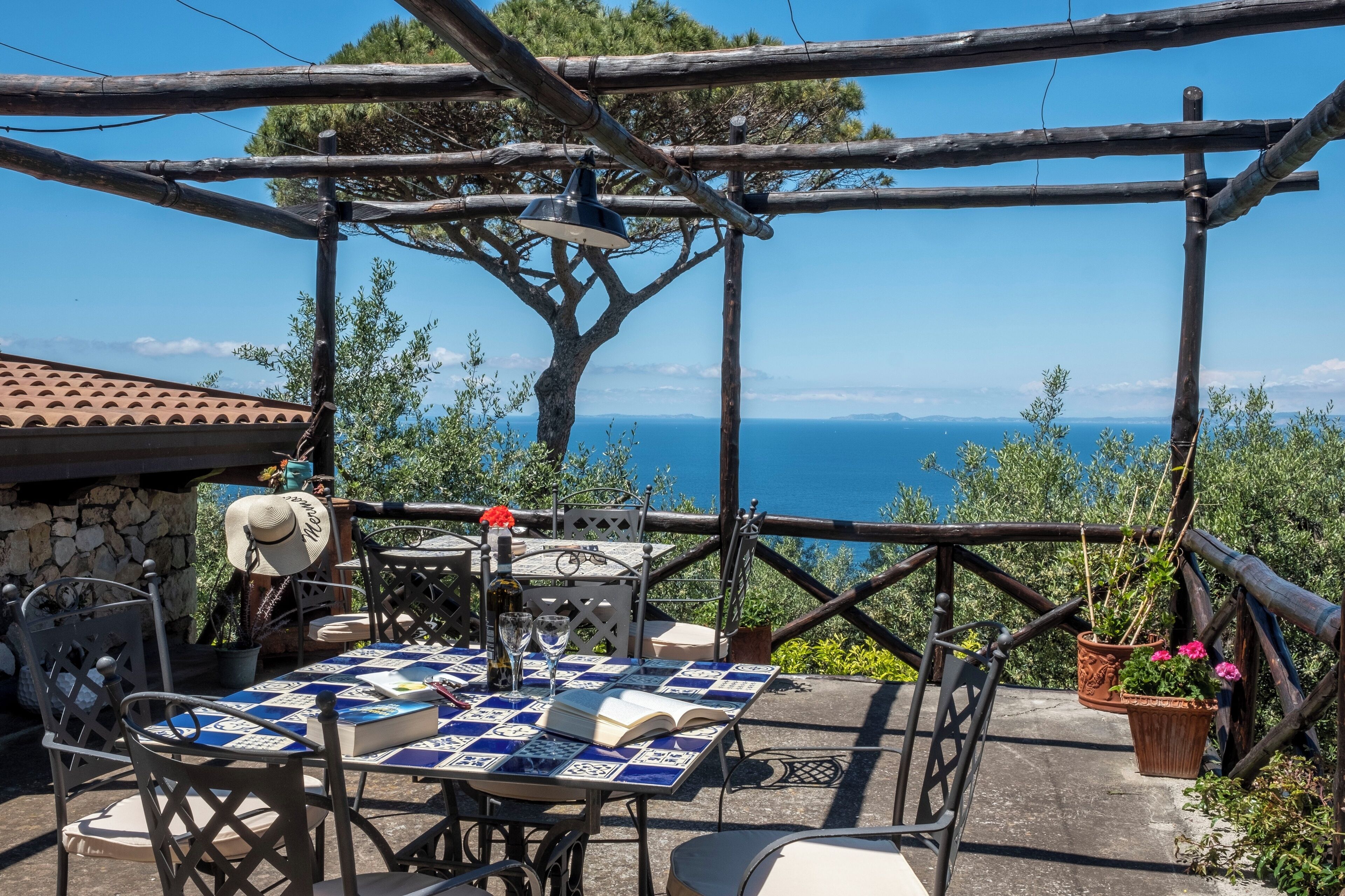 An outdoor dining area with a table set for two, surrounded by a wooden pergola, overlooking a scenic view of the sea.