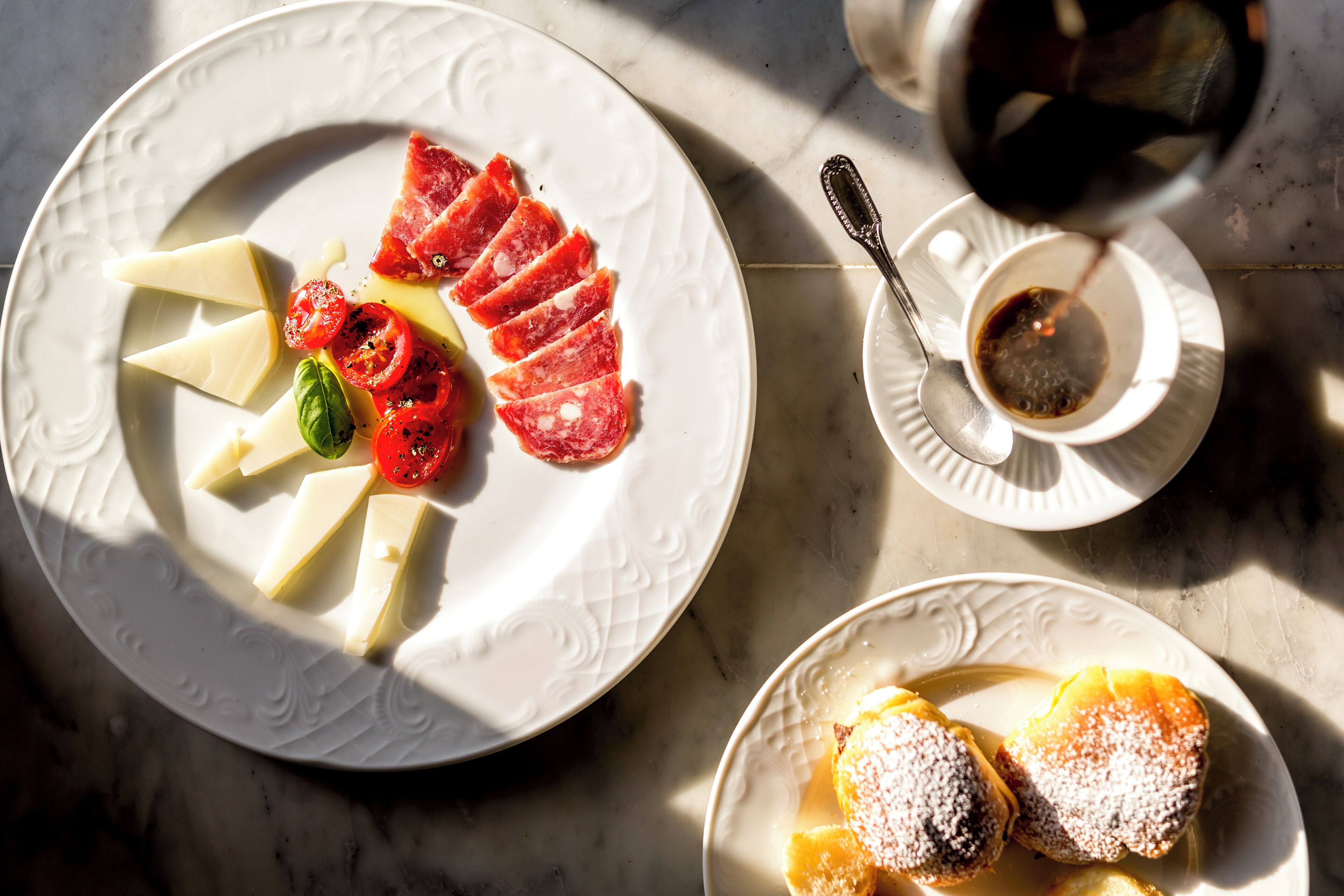 A plate with cheese, salami, and tomatoes, a cup of coffee, and a plate with pastries on a marble surface.