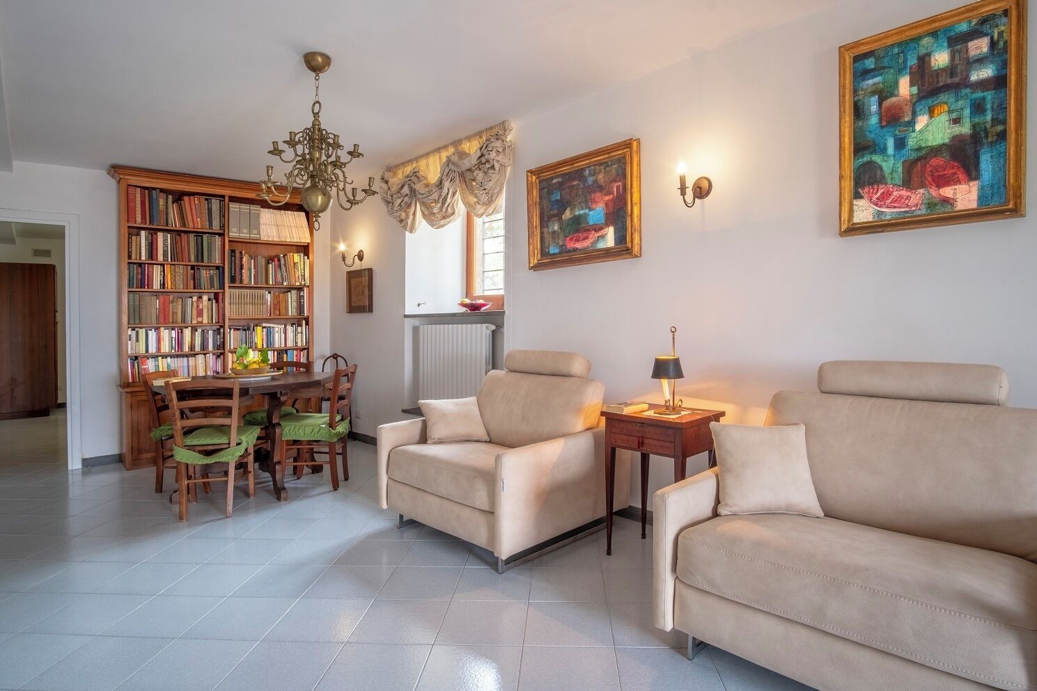 A living room with a bookshelf, two armchairs, a dining table with chairs, a chandelier, and two paintings.