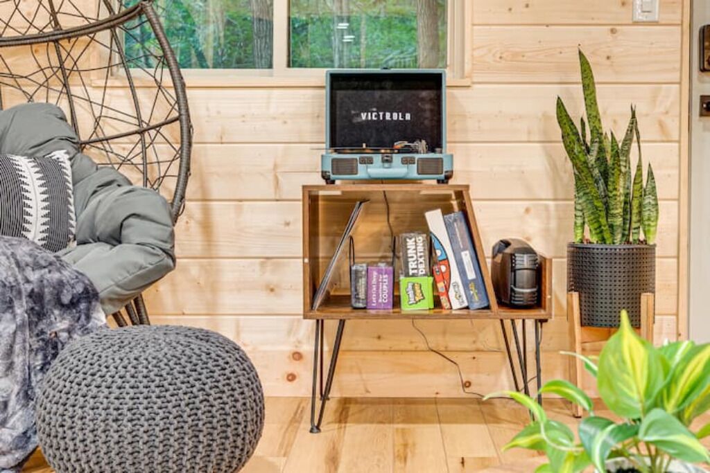 A cozy room with a wooden wall, a comfortable chair, a small wooden shelf with books and a coffee maker, and a window with a view of trees.