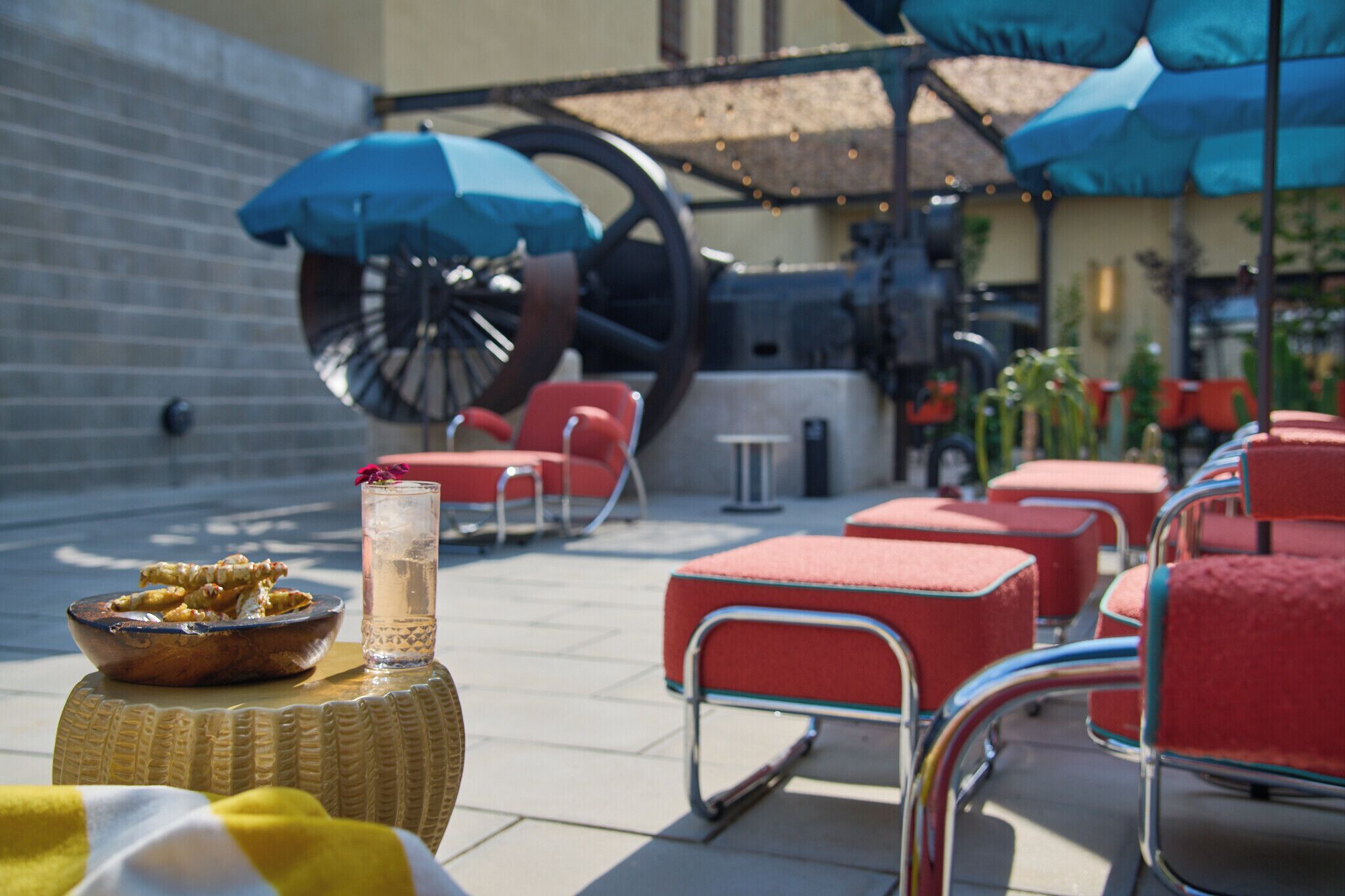 Un espace patio avec des chaises rembourrées rouges, une table en osier sur laquelle se trouve un saladier rempli de nourriture et une boisson, et des parasols bleus qui offrent de l’ombre.