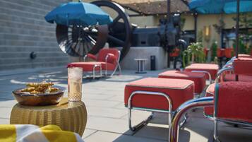 Un espace patio avec des chaises rembourrées rouges, une table en osier sur laquelle se trouve un saladier rempli de nourriture et une boisson, et des parasols bleus qui offrent de l’ombre.