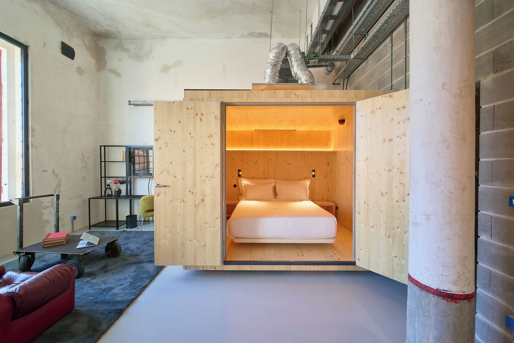 Quarto compacto e moderno, com uma cama, uma escrivaninha pequena e um sofá vermelho.