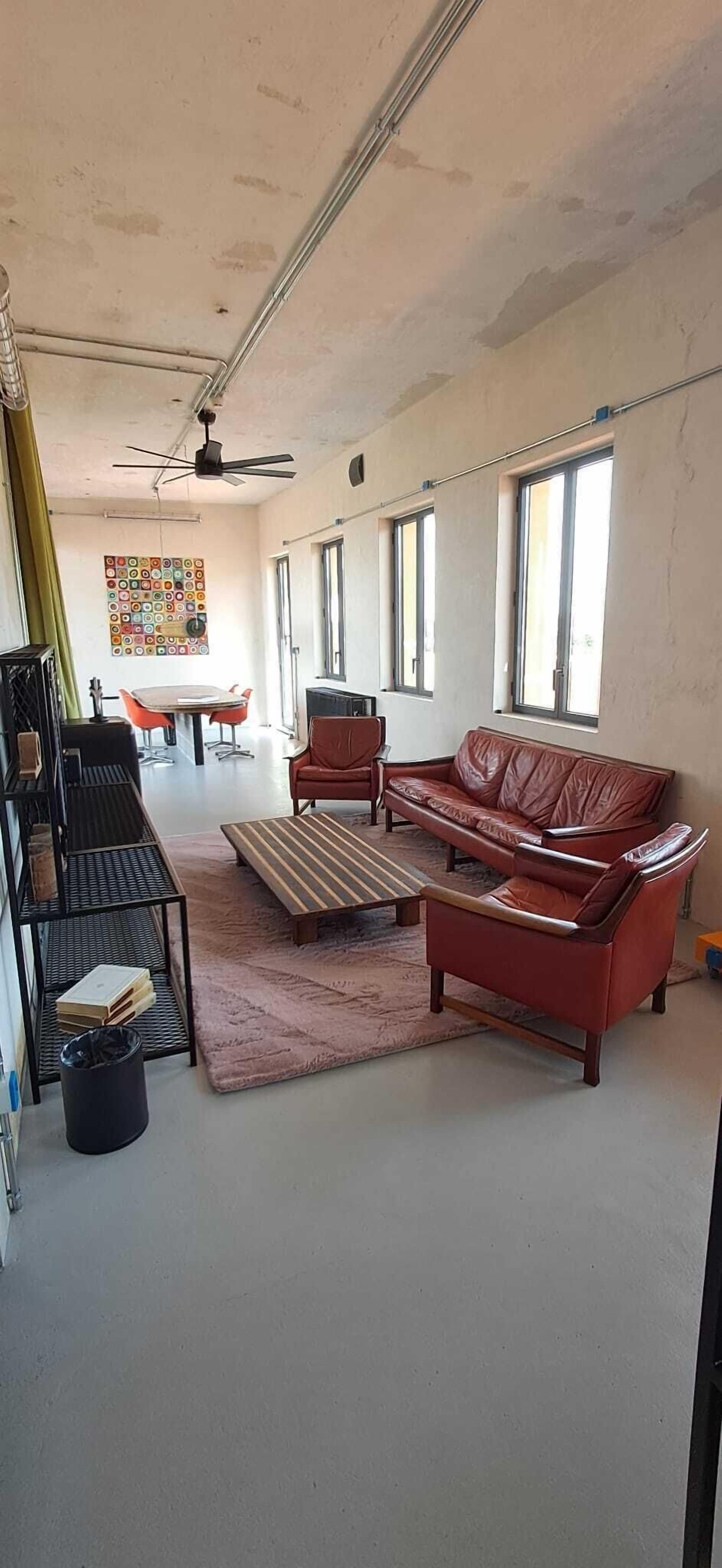 A spacious living area with a red leather sofa, a coffee table, and a dining table with chairs. There is a ceiling fan, large windows, and a colorful abstract painting on the wall.