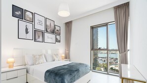 Un dormitorio moderno con una cama grande, una mesita de noche con lámpara, otra mesita auxiliar y vistas a un puerto deportivo a través de un ventanal amplio.