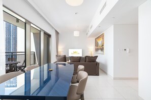 A modern hotel room with a glass dining table, a sofa, and a large window offering a city view.