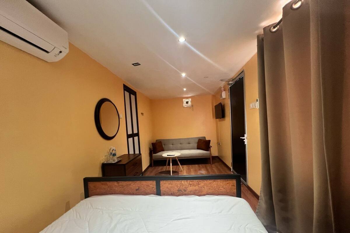 A hotel room with a bed, a sofa, a small table, and an air conditioning unit.