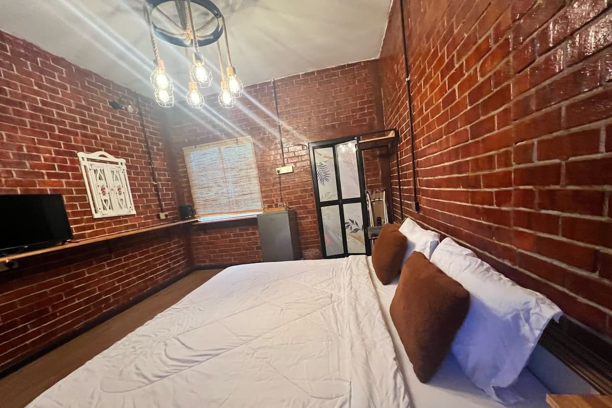 A hotel room with a brick wall, a bed with white bedding and brown pillows, a television on the left, and a door with a mirror and framed pictures.