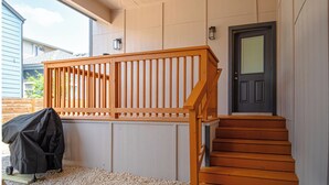 A wooden staircase leading to a dark door, a covered grill, and a view of a neighboring house.