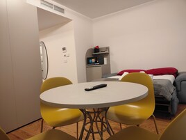 A small, modern room with a round table and yellow chairs, a bed with red and gray bedding, and a kitchenette area.