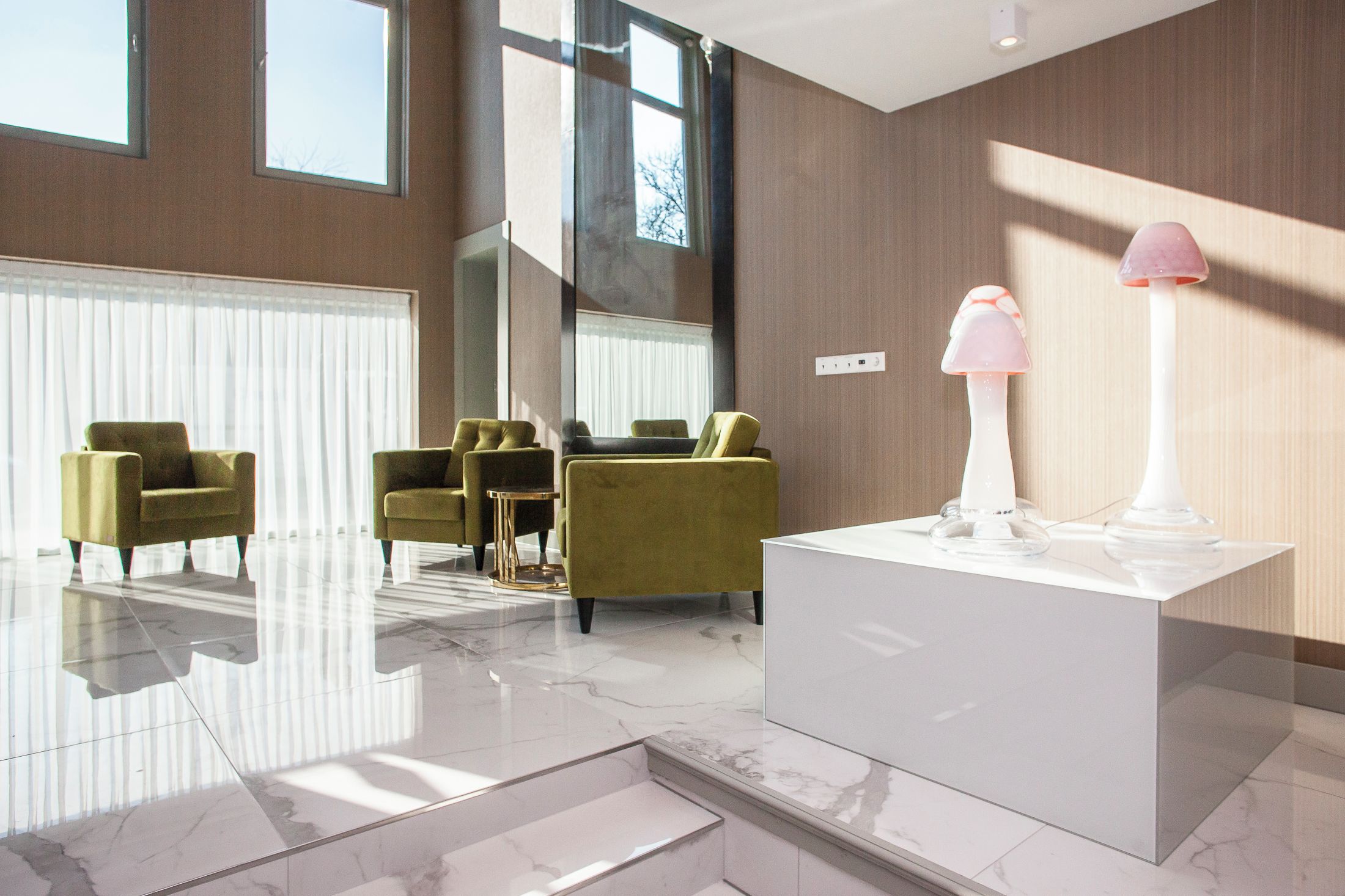 A modern lobby with a marble floor, green armchairs, a white reception desk, and a large window with blinds.