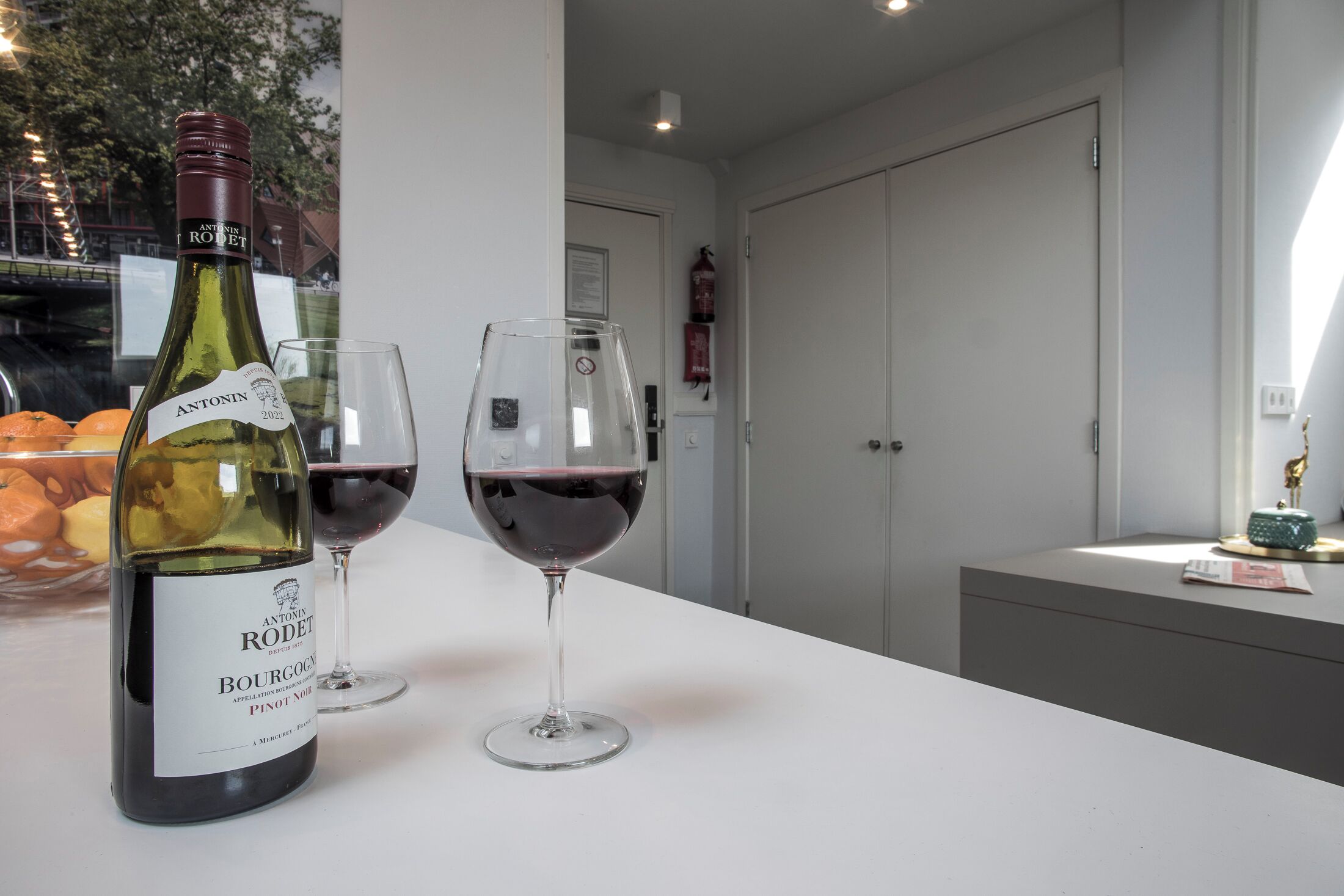 A bottle of Antonin Rodet wine and two glasses on a white table in a modern room with a view of a bridge and trees.
