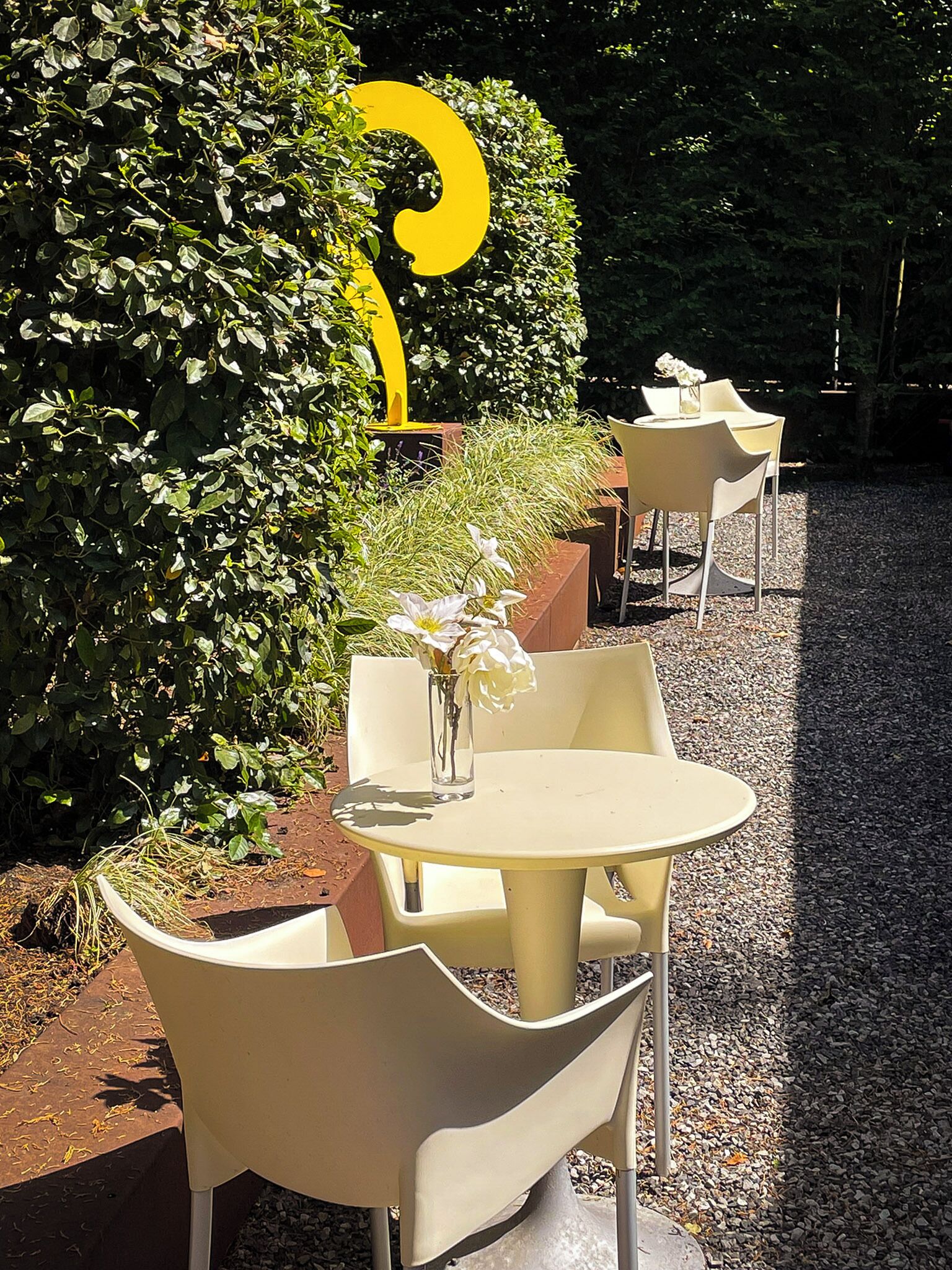 An outdoor seating area with round tables and chairs, surrounded by greenery and a large yellow letter 'G'.