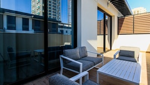 A modern outdoor seating area with a glass wall, a wooden deck, and a view of a high-rise building. - Modern 1BR Living in Oakley Square (Dubai)