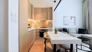 A modern kitchen with built-in appliances, a dining area with a table and chairs, and a living area with a sofa and a plant. - Modern 1BR Living in Oakley Square (Dubai)
