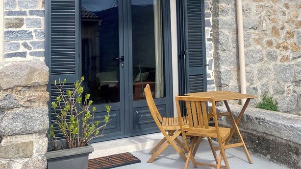A stone facade with a balcony, a sliding glass door, and a wooden table and chair set outside. - Small village house (Bastelica)