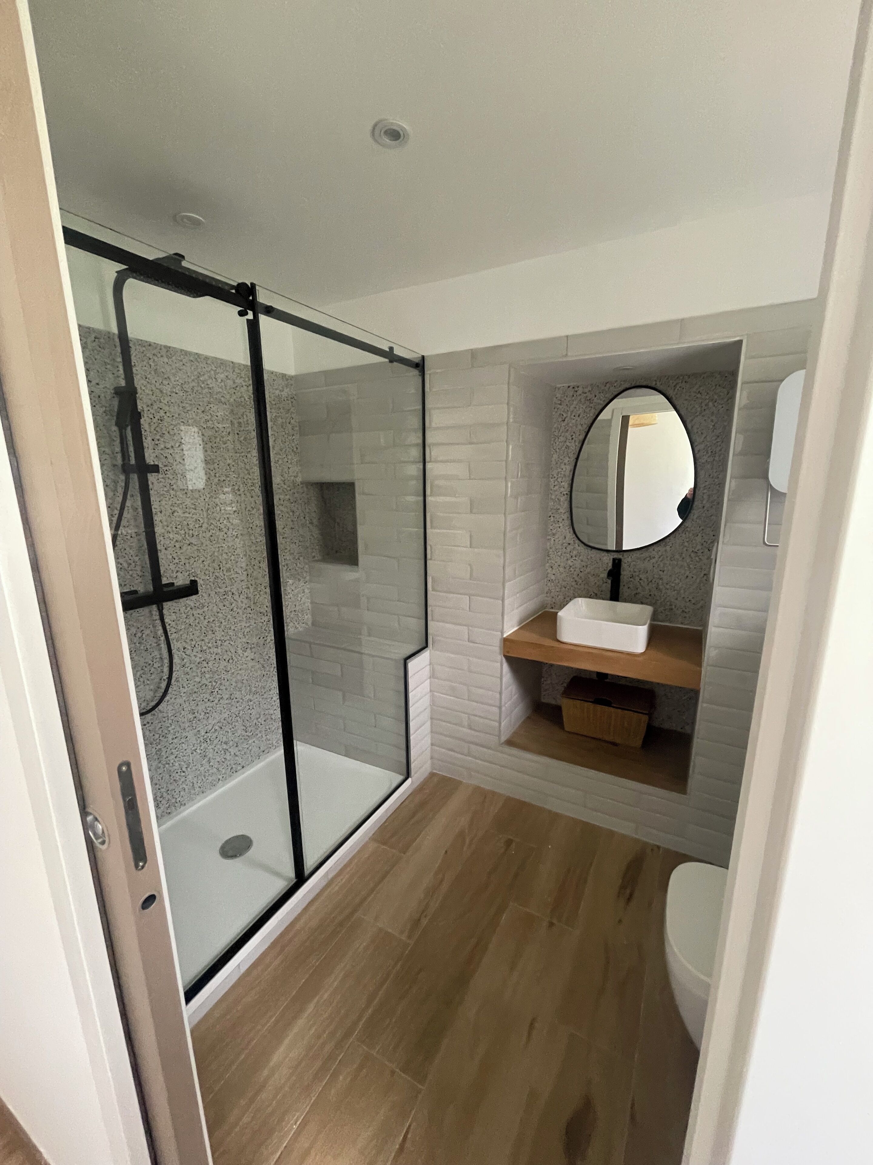 A modern bathroom with a glass-enclosed shower, a white sink with a mirror above it, and a wooden shelf.
