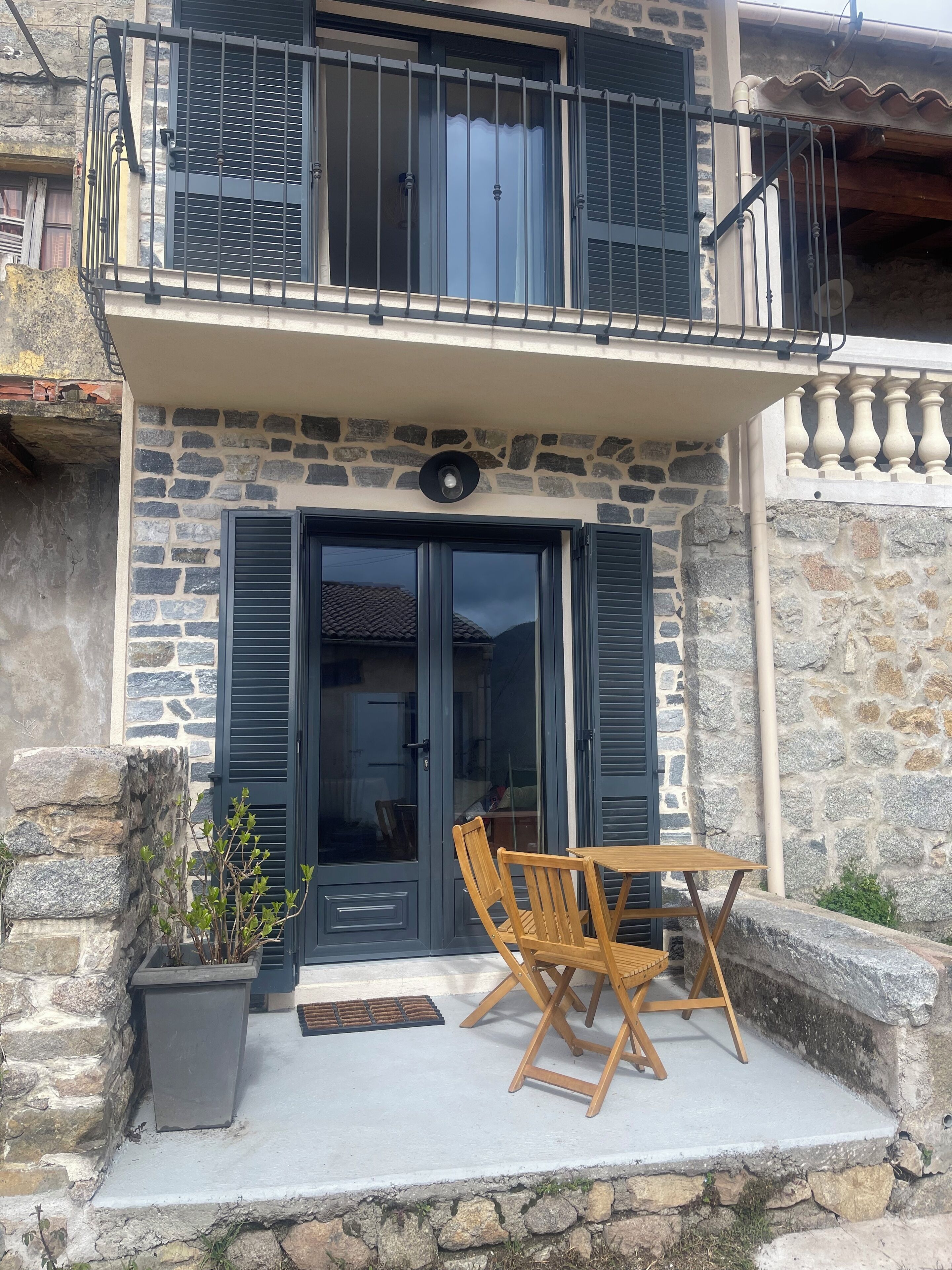 A stone building with a balcony, a wooden table and chairs, and a potted plant.