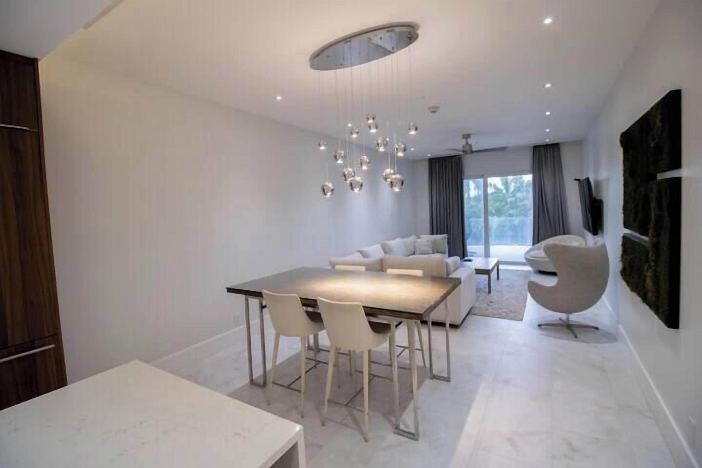 A modern dining area with a white table and chairs, a contemporary chandelier, and a sofa in the background.