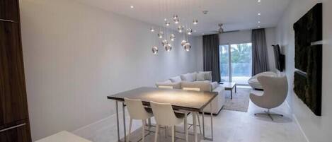 A modern dining area with a white table and chairs, a contemporary chandelier, and a sofa in the background.