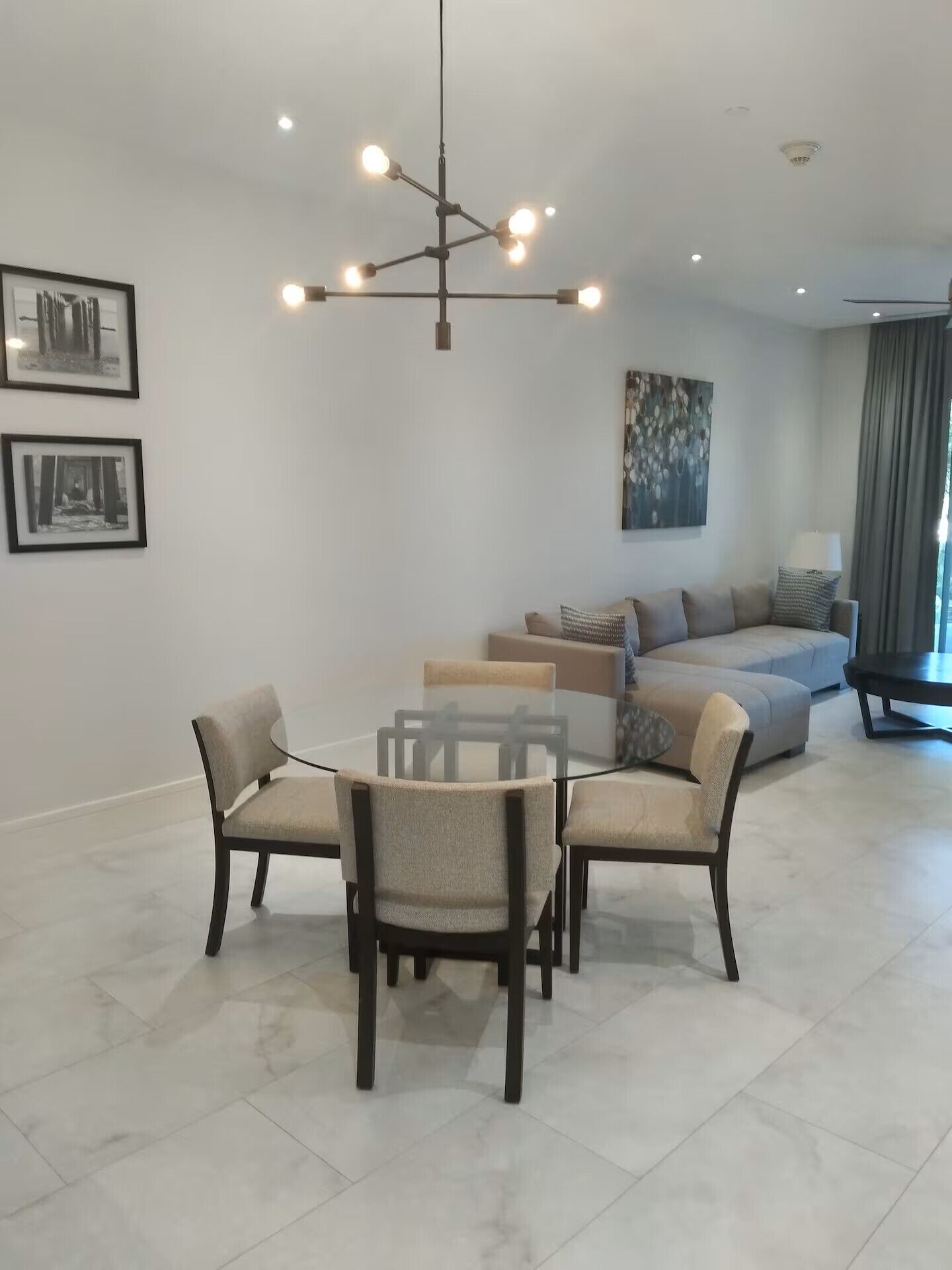 Uma sala de estar moderna com uma mesa de jantar redonda, quatro cadeiras, um sofá seccional e um lustre contemporâneo.