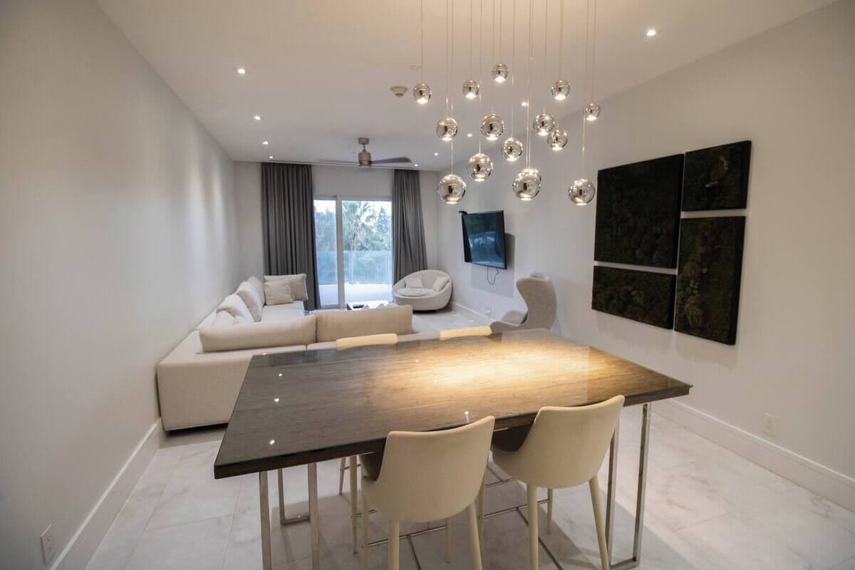 A modern living room with a dark wooden dining table, beige sofas, and a large window with curtains.