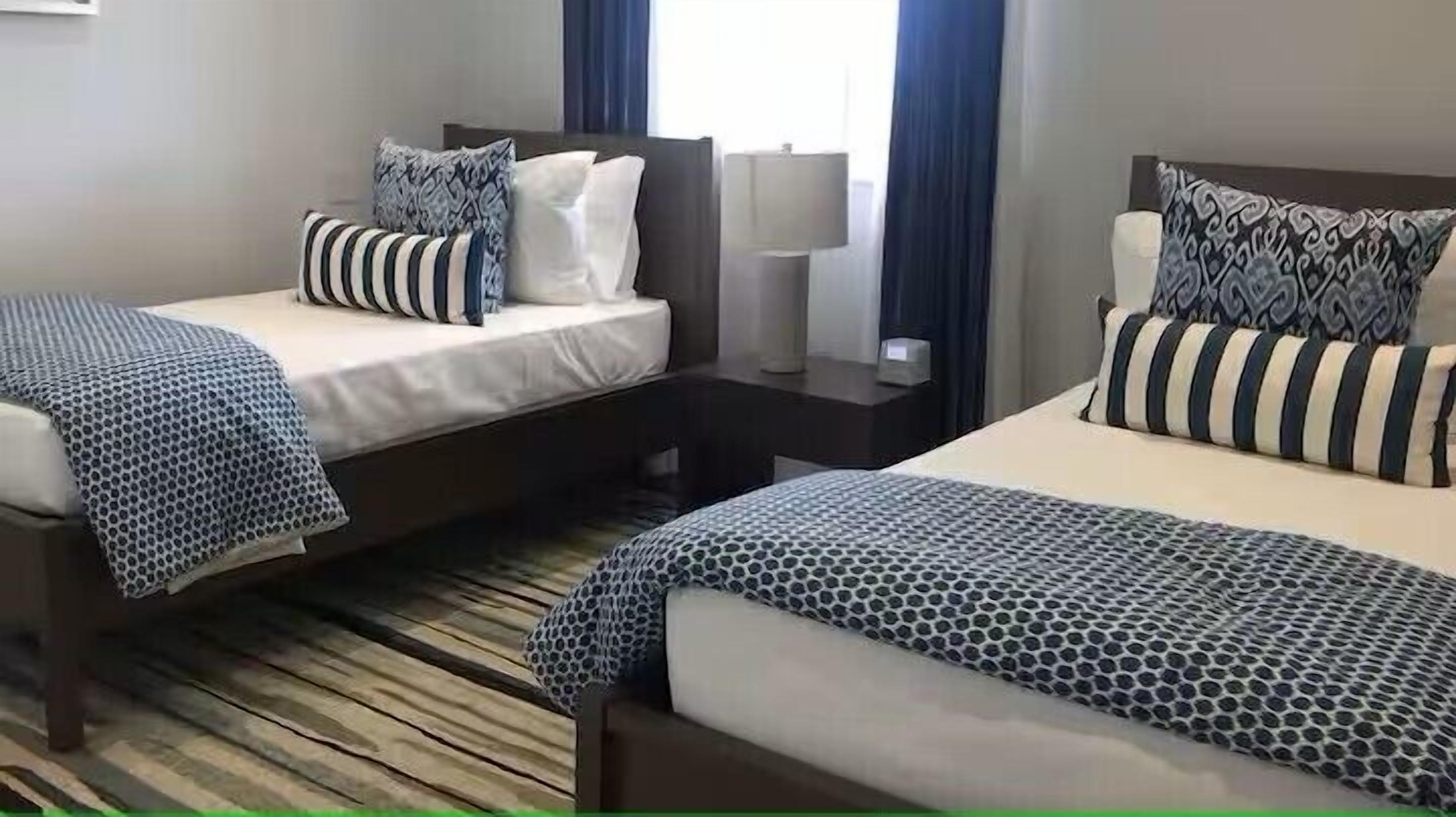 Una habitación de hotel con dos camas, cada una con una colcha estampada y almohadas a juego. Hay una mesita de noche con una lámpara entre las camas, y una ventana con cortinas.