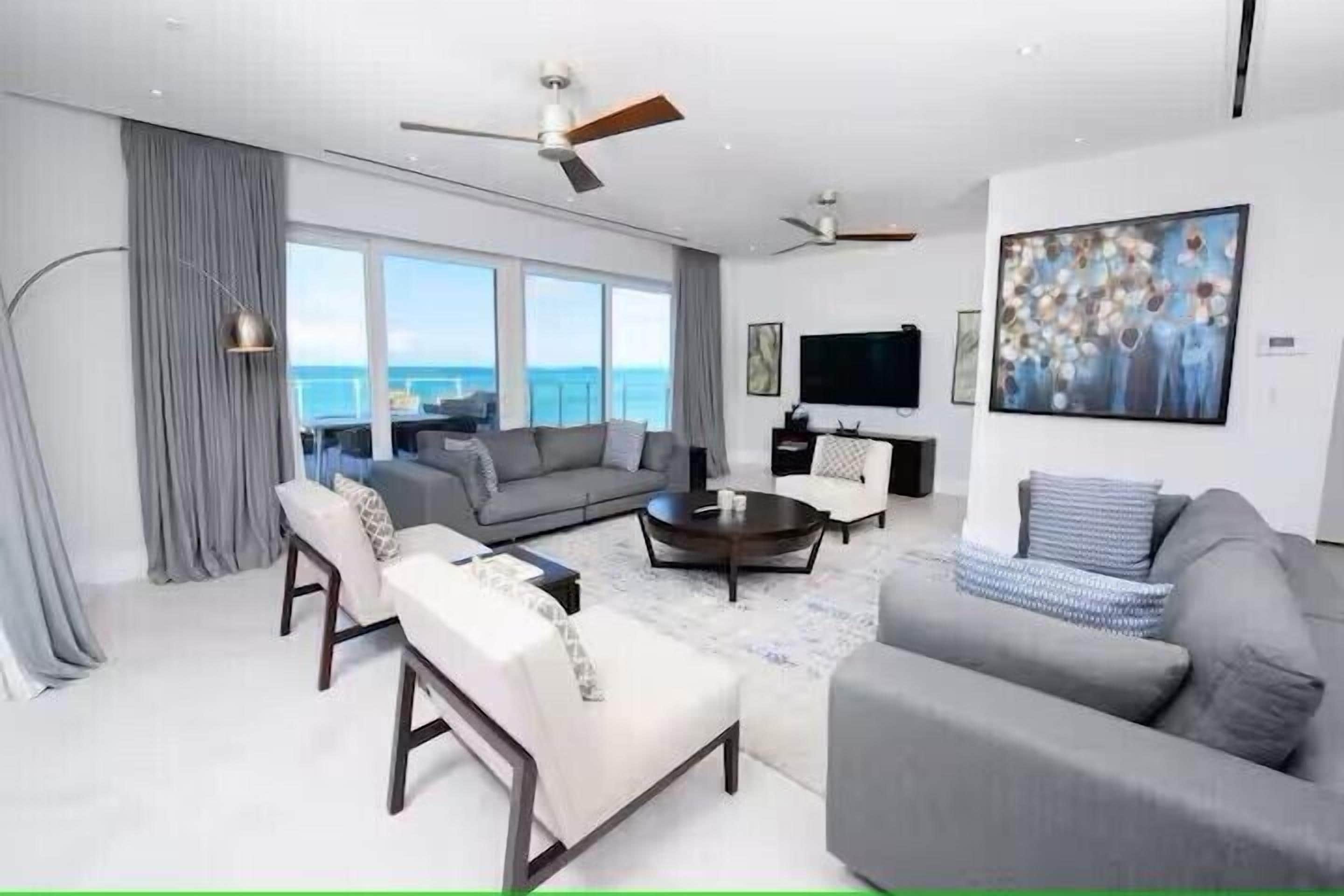 A modern living room with a sofa set, a coffee table, and a flat-screen TV. There is a large window with a view of the ocean.