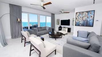 A modern living room with a sofa set, a coffee table, and a flat-screen TV. There is a large window with a view of the ocean.