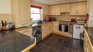 A modern kitchen with wooden cabinets, a built-in washing machine, a microwave, and a small oven. - 6 Wildersmouth Court Apartments (Ilfracombe)