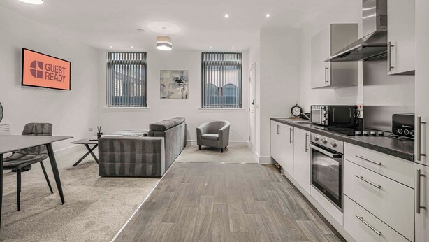 Una cocina moderna con armarios blancos, encimera negra y electrodomésticos de acero inoxidable. Cuenta con una zona de comedor con mesa y sillas, y un rincón de descanso con sofá y sillón.