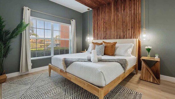 A bedroom with a wooden bed frame, a large window with curtains, a wooden slatted ceiling, and a bedside table with a vase.