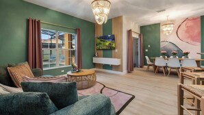 A modern living room with a green wall, a large window, a dining area with a table and chairs, and a chandelier.