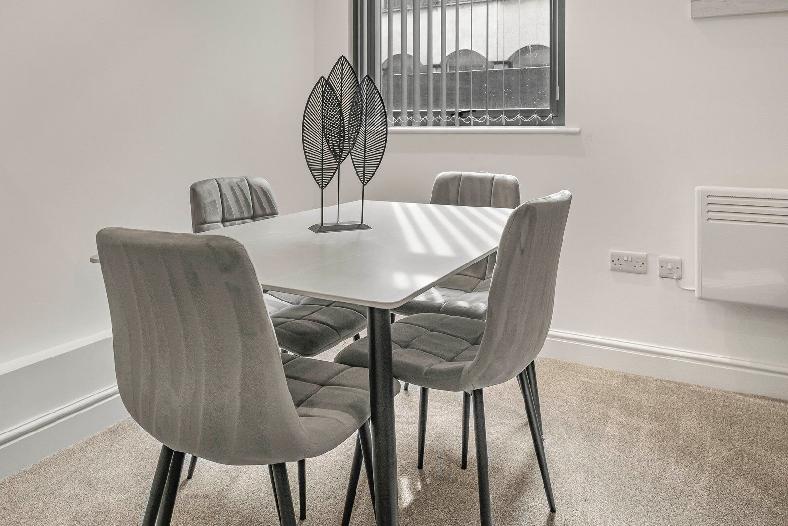 A modern dining area with a white table and four grey upholstered chairs. There is a decorative black metal piece on the table, and a window with bars is visible in the background.