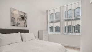 Un dormitorio minimalista con una cama grande, una mesita de noche, un aparador y una ventana con vistas a edificios.