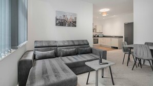 Una sala moderna con un sofá seccional gris, una mesita de centro y un comedor con mesa y sillas. Se ve una cocina al fondo.