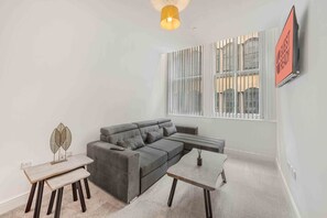 Un salon moderne avec un canapé d’angle gris, une petite table et un objet décoratif en métal posé sur le sol.