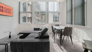 Quarto de hotel moderno com sofá cinza, uma mesa de jantar com cadeiras, janelas amplas com persianas verticais e um aviso que diz 'GUEST READY'.