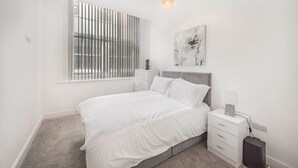 A bedroom with a bed, a nightstand, a lamp, a power outlet, a window with blinds, and a ceiling light.