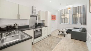 A modern kitchen with a built-in oven, a microwave, and a kettle. There is a dining area with a table and chairs, and a sofa.