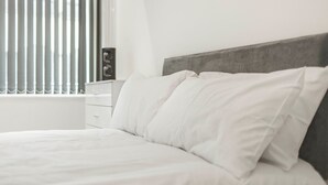 A neatly made bed with white bedding and pillows, a white nightstand with a small item on it, and a window with blinds.