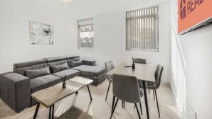 A modern living room with a grey sectional sofa, a small round table, and a dining table with chairs. There are two windows with blinds, a ceiling light, and a 'GUEST READY' sign on the wall. - GuestReady - Relaxing retreat in Liverpool (Liverpool)