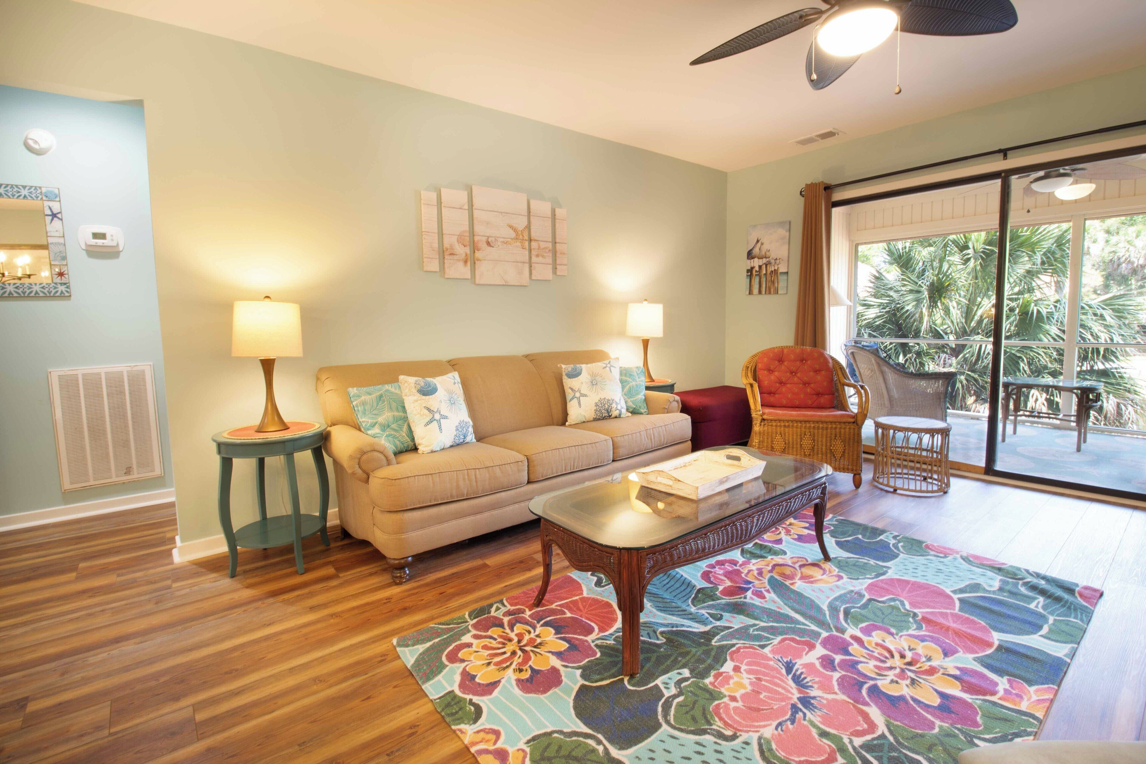 Un salon avec un canapé beige, deux tables d’appoint avec des lampes, une table basse et un grand tapis à motifs floraux. Une porte-fenêtre coulissante donne sur un espace extérieur aménagé avec une chaise et une table.