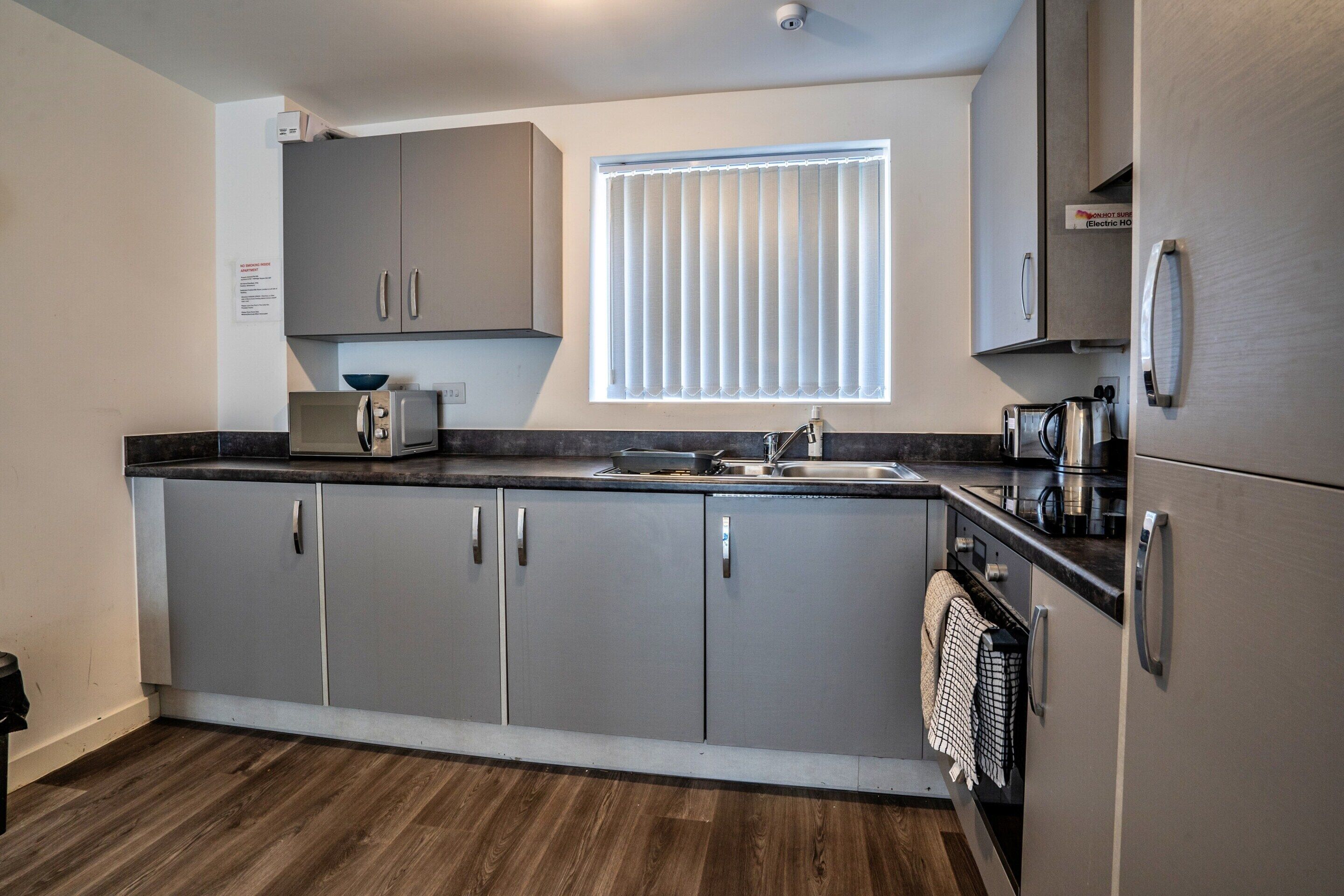 A modern kitchen with grey cabinets, a built-in oven, a microwave, and a window with blinds.