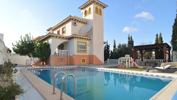 A swimming pool with a sun terrace, a covered seating area, and a tall building with a tower. - Villa Amore Mio South (Orihuela Costa)