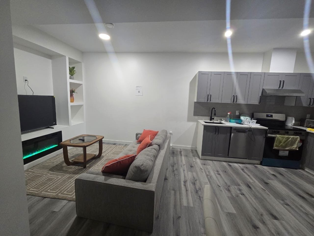 A modern living room with a sofa, a coffee table, and a TV. There is a kitchen area with cabinets and appliances.
