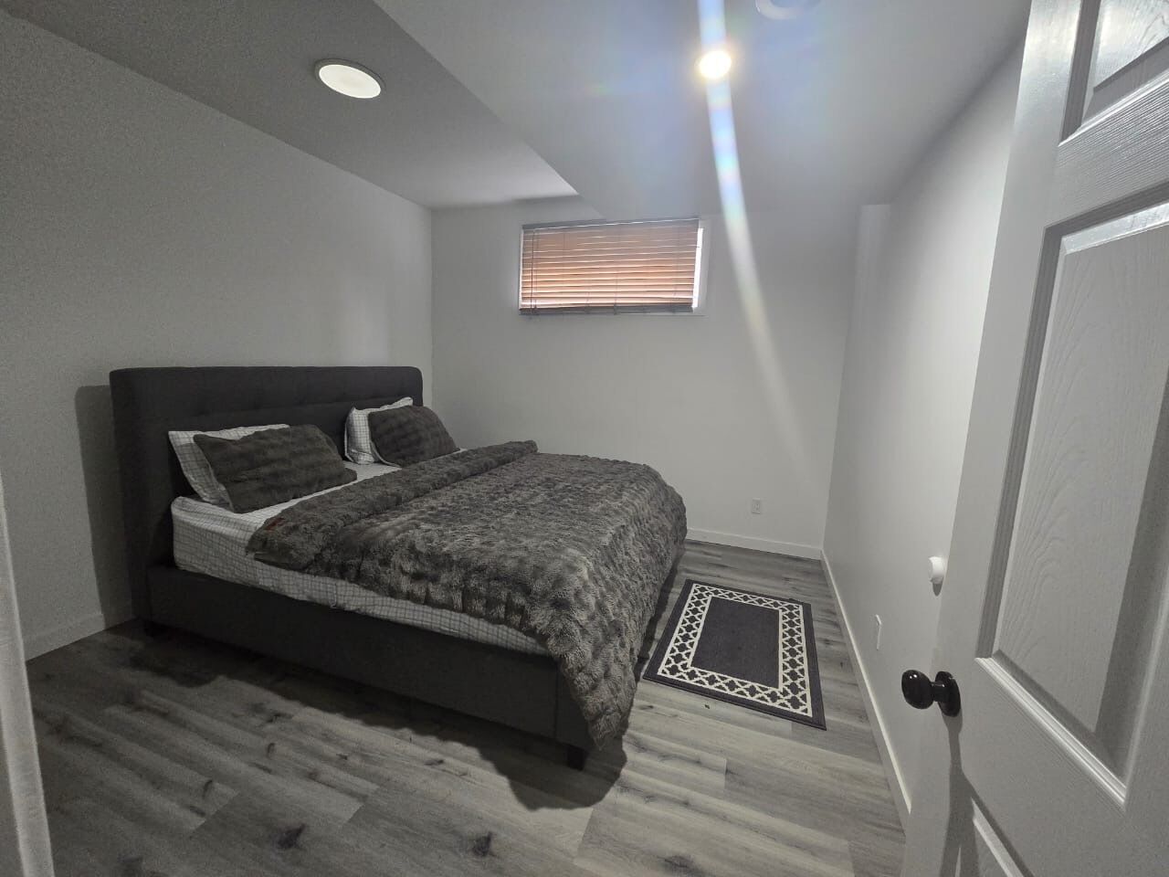 A bedroom with a bed, a window with blinds, a door, and a small rug.